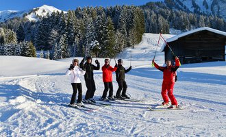 A group of five skiers stands happily in the snow, enjoying the sunny day. In the background, there are snow-covered trees and a wooden house. | © Andy Herr | Werbewind GmbH