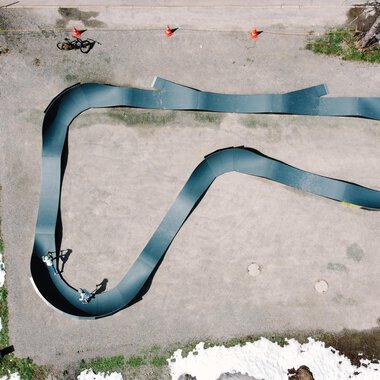 Eine Draufsicht auf eine geschwungene Rennbahn, die aus schwarzem Material besteht. Rund um die Bahn sind einige Menschen und Schneereste zu sehen. | © Christian Posch | Klaus Zopf