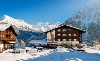 Eine charmante Alpenlandschaft im Winter mit schneebedeckten Bergen. Im Vordergrund befindet sich ein Hotel und mehrere parkende Autos.