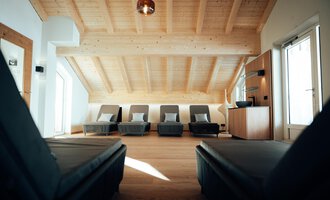 A modern room with a wooden ceiling and loungers. There are large windows that let in plenty of light.