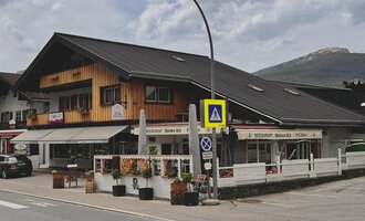Ein traditionelles alpenländisches Gebäude mit Holzverkleidung und einem Restaurant im Erdgeschoss. Im Vordergrund sind Blumenarrangements und Verkehrssignale zu sehen. | © Riezlern Eck | Sead Sulejmani
