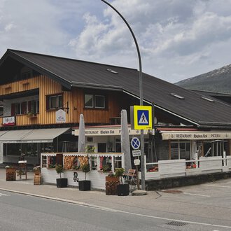 A traditional alpine building with wooden cladding and a restaurant on the ground floor. In the foreground, flower arrangements and traffic signs can be seen. | © Riezlern Eck | Sead Sulejmani