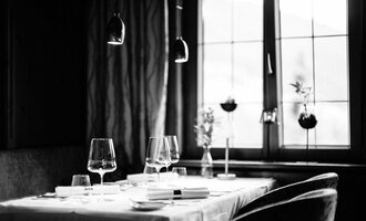 Ein eleganter Restauranttisch mit stilvollen Gläsern und einem weißen Tischtuch. Die Fenster im Hintergrund lassen sanftes Licht herein und zeigen Pflanzendekoration.