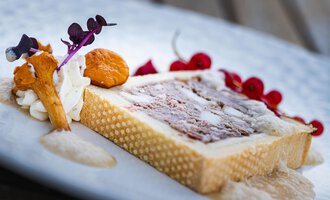 Ein kunstvoll angerichtetes Dessert auf einem weißen Teller. Es besteht aus einer rechteckigen Form, garniert mit essbaren Blumen und Früchten.