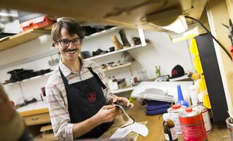 Ein Schuhmacher in seiner Werkstatt lächelt, während er an einem Schuh arbeitet. Im Hintergrund sind verschiedene Werkzeuge und Materialien zu sehen. | © Kleinwalsertal Tourismus | Oliver Farys