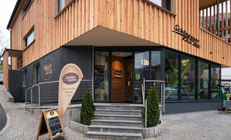 Ein modernes Restaurant mit Holzfassade und großen Fenstern. Der Eingangsbereich ist einladend gestaltet, mit einem Schild für das Restaurant. | © Schönegger Käse-Alm