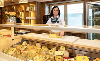 A cheese seller stands behind a sales counter with a large selection of cheese. The shop has a bright and inviting atmosphere. | © Schönegger Käse-Alm