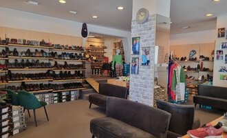 A shoe store with shelves full of shoes and a cozy seating area. Pictures hang on the walls and some clothing items are visible. | © Schuhhaus Schuster | Marietta Diehl