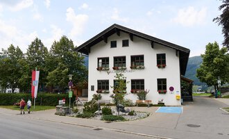 Ein charmantes, traditionelles Gebäude mit einem Walmdach. Es ist von Bäumen umgeben und hat blühende Fensterkästen. | © Kleinwalsertal Tourismus | Oliver Farys
