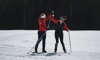 Two women in warm sportswear high-five each other while cross-country skiing. They are standing on a snow-covered area in a beautiful winter landscape.
