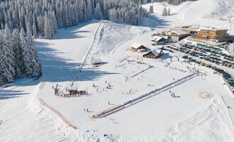 Ein schneebedeckter Skiort mit Pistenfahrern und einem Skilift. Im Hintergrund sind sich sanft neigende Hügel und gemütliche Hütten zu sehen.