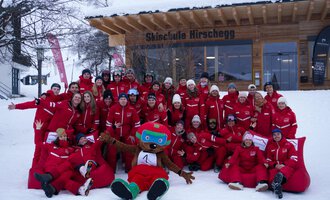 Eine Gruppe von Skilehrern in roten Anzügen steht vor einer Skischule im Schnee. Ein Maskottchen in einem bunten Kostüm sitzt im Vordergrund und lächelt.