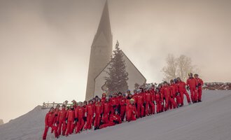 A group of people in red ski suits is standing in the snow in front of a church. The landscape is wintry and surrounded by fog. | © Skischule Mittelberg | Karl Schuster