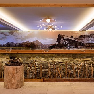 An elegant interior with a stone wall and a large mirror. In the background, there is a large landscape painting featuring a cabin and mountains. | © Aparthotel Kleinwalsertal Haller GmbH & Co KG