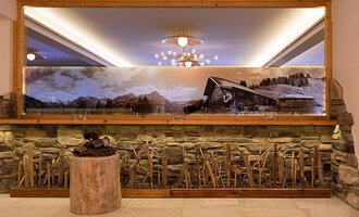 An elegant interior with a stone wall and a large mirror. In the background, there is a large landscape painting featuring a cabin and mountains. | © Aparthotel Kleinwalsertal Haller GmbH & Co KG