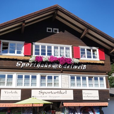 A traditional wooden house with red shutters and floral decorations. The clear sky and the surrounding mountains create an idyllic atmosphere. | © Sport Edelweiß | N. Lughammer