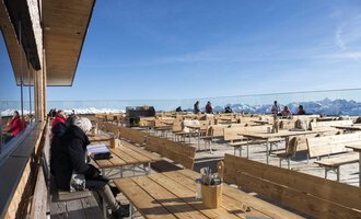 An idyllic restaurant on a mountain terrace with a variety of wooden tables and chairs. In the background, snow-capped mountains and a clear sky can be seen. | © OBERSTDORF · KLEINWALSERTAL BERGBAHNEN