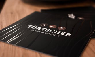 An elegant menu booklet with the inscription "TÜRTSCHER – the joiner's workshop". It features various wood symbols on a dark background. | © Türtscher Die Schreinermanufaktur