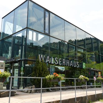 Ein modernes Gebäude mit großen Glasfenstern, umgeben von grünen Pflanzen. Es trägt die Beschriftung "WALSERHAUS". | © Kleinwalsertal Tourismus | Oliver Farys