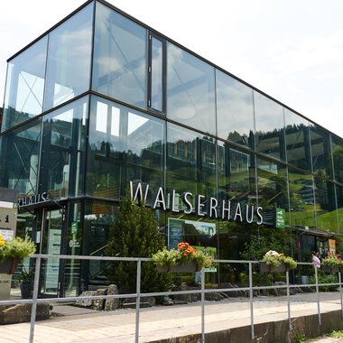 Ein modernes Gebäude mit großen Glasfenstern, umgeben von grünen Pflanzen. Es trägt die Beschriftung "WALSERHAUS". | © Kleinwalsertal Tourismus | Oliver Farys