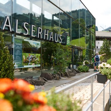 The Walserhaus is a modern building with large windows in a picturesque setting. In the foreground, colorful flowers and a hiker can be seen. | © Kleinwalsertal Tourismus | Oliver Farys