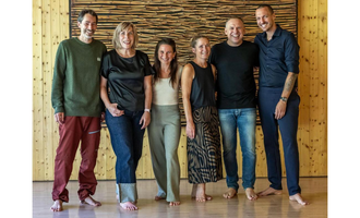 Six people are smiling together in front of a wall clad in wood.  
They are all dressed casually and are standing barefoot on a light floor.