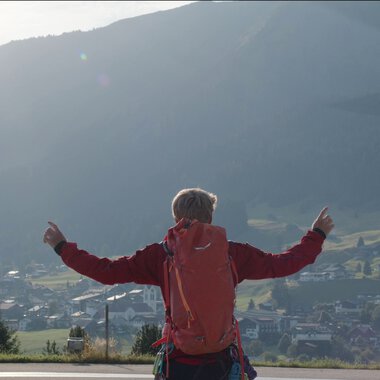 Eine Person mit rotem Rucksack steht auf einer Wiese und zeigt nach oben, während ein Hubschrauber im Flug über der Landschaft fliegt. | © Kleinwalsertal Tourismus eGen