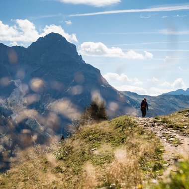 A hiker walking on a trail in the mountains of the Black Forest on a sunny day, surrounded by green grass and mountain peaks in the background. | © Kleinwalsertal Tourismus eGen • Photographer: Andre Tappe