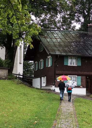 Zwei Personen mit Regenschirmen gehen auf einem Gehweg an einem Haus in einer grünen Landschaft vorbei. | © Kleinwalsertal Tourismus eGen • Fotograf: Andre Tappe
