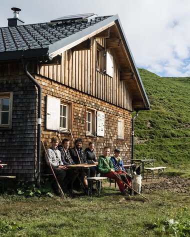 Eine Gruppe von Menschen sitzt draußen vor einem alten Holzhaus in einer grünen, bergigen Landschaft. | © Kleinwalsertal Tourismus eGen • Fotograf: Andre Tappe