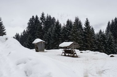 Schneelandschaft mit Holzhäuschen und einem Unterschlupf in einem Waldgebiet, verschneit und winterlich. | © Kleinwalsertal Tourismus eGen • Fotograf: Andre Tappe