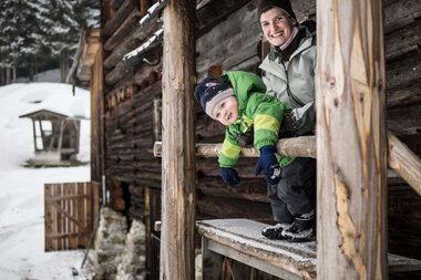 Eine Frau und ein Kind schauen aus einem Fenster eines Holzhauses im Schnee, lächeln und genießen die winterliche Natur. | © Kleinwalsertal Tourismus eGen • Fotograf: Andre Tappe