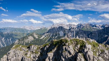 Mountain summit with cross and hikers, above the Alps, on a clear day with blue sky and clouds. | © Kleinwalsertal Tourismus eGen • Photographer: Bastian Morell