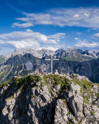 Mountain summit with cross and hikers, above the Alps, on a clear day with blue sky and clouds. | © Kleinwalsertal Tourismus eGen • Photographer: Bastian Morell