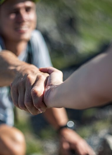 Two hikers reaching out and holding hands, symbolizing support during a mountain hike. | © Kleinwalsertal Tourismus eGen • Photographer: Bastian Morell