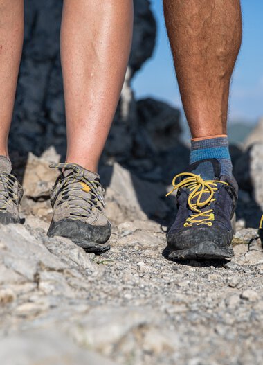 Close-up of three hiking feet on a rocky trail, wearing different colored hiking shoes. | © Kleinwalsertal Tourismus eGen • Photographer: Bastian Morell