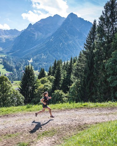 A person running on a trail in a lush green mountain landscape with tall mountains in the background. | © Kleinwalsertal Tourismus eGen • Photographer: Dominik Berchtold