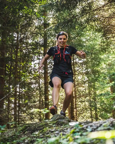 Woman trail running in a dense forest, happily jumping over a tree trunk. | © Kleinwalsertal Tourismus eGen • Photographer: Dominik Berchtold