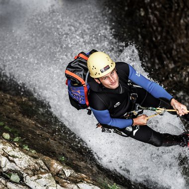 Adventurer rappelling down a waterfall, wearing a helmet and full gear, holding onto a rope in a rugged outdoor setting. | © Kleinwalsertal Tourismus eGen • Photographer: Dominik Berchtold