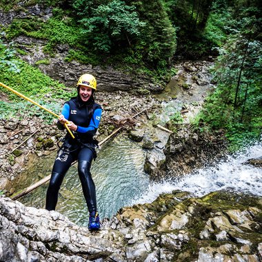 Woman canyoning in a river surrounded by lush green forest, wearing a wetsuit, helmet, and secured with a rope. | ©   Kleinwalsertal Tourismus eGen • Photographer: Dominik Berchtold