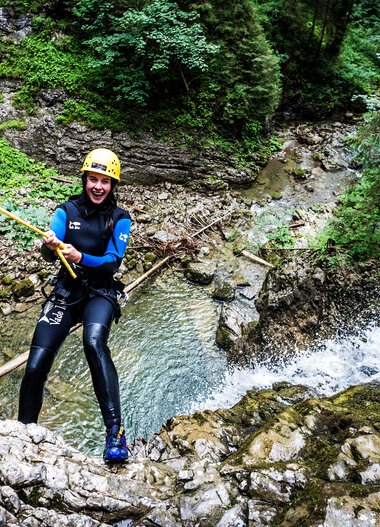 Frau beim Canyoning in einem Fluss inmitten eines grünen Waldes, trägt Neoprenanzug, Helm und ist an einem Seil befestigt. | © Kleinwalsertal Tourismus eGen • Fotograf: Dominik Berchtold