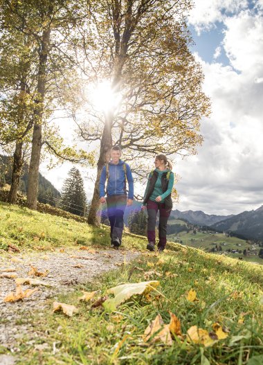 Zwei Wanderer gehen den Wanderweg in einer Herbstlandschaft mit Bergen, Bäumen und wolkigem Himmel, die Sonne scheint durch die Bäume. | © Kleinwalsertal Tourismus eGen • Fotograf: Dominik Berchtold