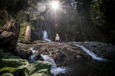 Person sitzt auf Felsen im Schwarzwasserbach in einem Wald, Sonnenlicht scheint durch die Bäume, Wasser plätschert um sie herum. | © Kleinwalsertal Tourismus eGen • Fotograf: Dominik Berchtold