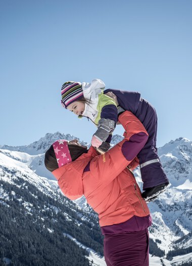 Family winter hiking in the mountains, an adult lifts a child onto a snow mound, with snow-covered peaks under a blue sky in the background. | © Kleinwalsertal Tourismus eGen • Photographer: Dominik Berchtold
