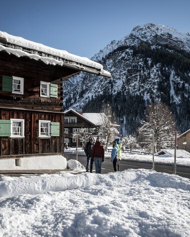 Drei Personen wandern auf einer verschneiten Straße in einem winterlichen Dorf, mit Bergen im Hintergrund. | © Kleinwalsertal Tourismus eGen • Fotograf: Dominik Berchtold