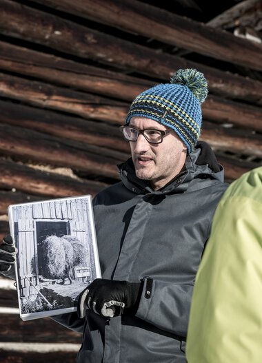 Ein Mann erklärt eine historische Fotografie vor einer Gruppe bei einer Kulturführung im Winter. | © Kleinwalsertal Tourismus eGen • Fotograf: Dominik Berchtold