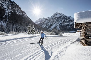 Cross-country skier on a snowy trail in a winter landscape with mountains and the sun in the background. | © Kleinwalsertal Tourismus eGen • Photographer: Dominik Berchtold