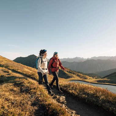 Two hikers walking on a narrow trail in the mountains with a lake in the background, sunny weather, panoramic mountain landscape. | © Kleinwalsertal Tourismus eGen • Photographer: Hannah Bichay
