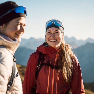 Two smiling women hiking in the mountains, dressed in outdoor gear and sunglasses.