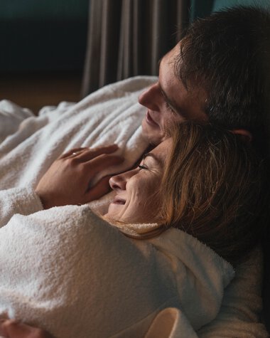 A happy couple is lying in bed, with the man embracing the woman, both smiling and appearing relaxed and in love. | © Kleinwalsertal Tourismus eGen • Photographer: Hannah Bichay